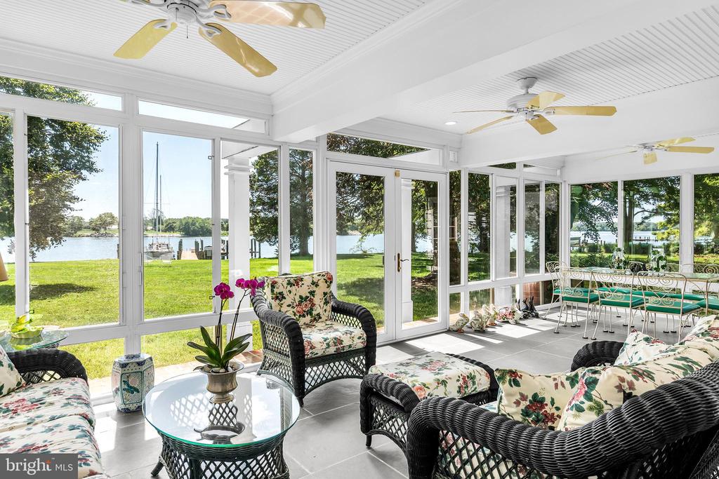 Furnished sunroom with floral wicker seating and broad windows overlooking a lawn and waterfront.