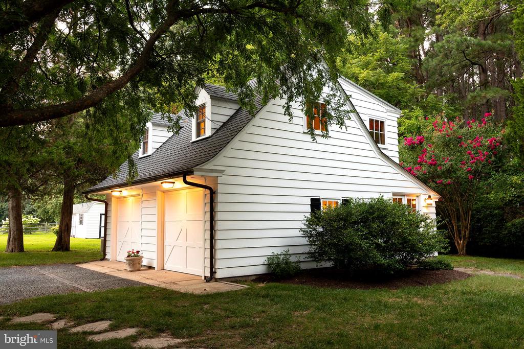 White two-story carriage house with double garage doors, dormer windows, and mature trees framing the lawn.