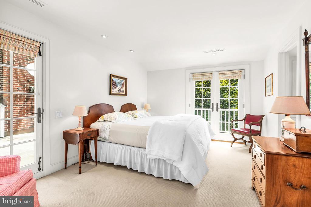 Bright bedroom with white bedding, wooden furniture, and French doors overlooking greenery.