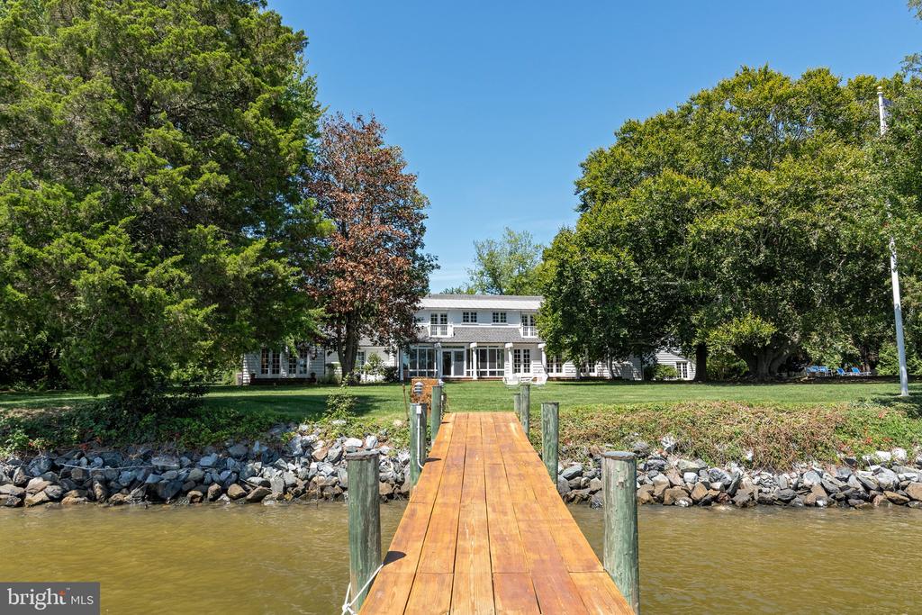 White two-story waterfront house framed by mature trees, viewed from a wooden dock.