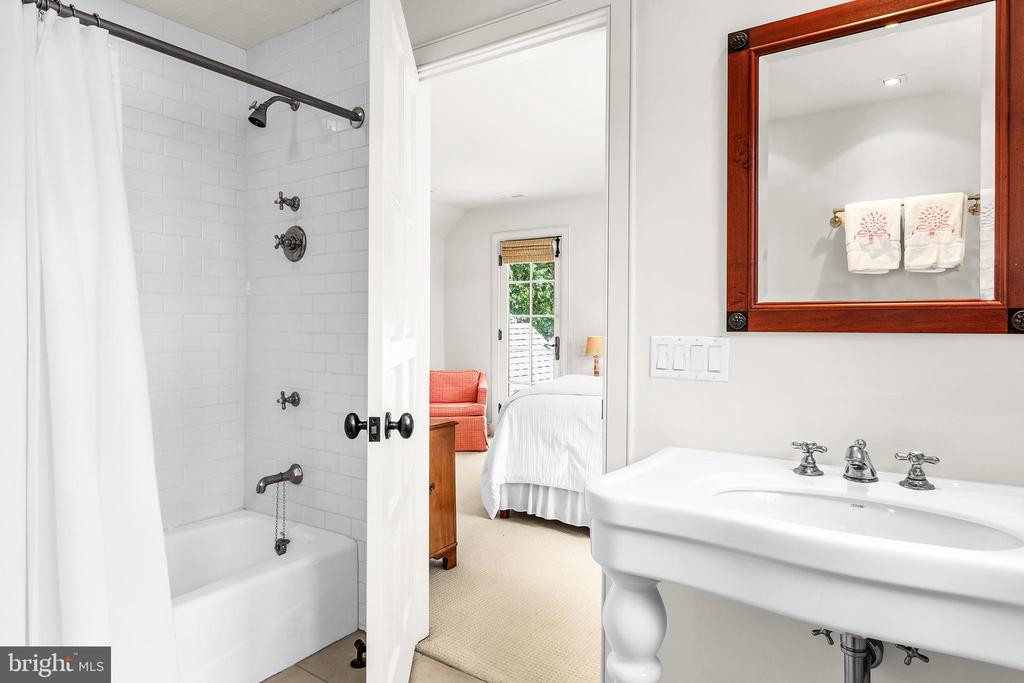 Bathroom with a subway-tile tub, pedestal sink, and wood-framed mirror beside an open bedroom doorway.