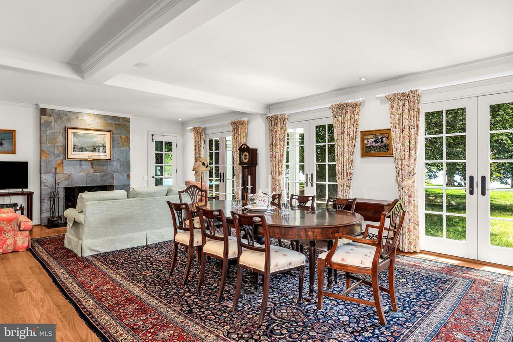 Dining and living room with a round wooden table, stone fireplace, and French doors overlooking the lawn.