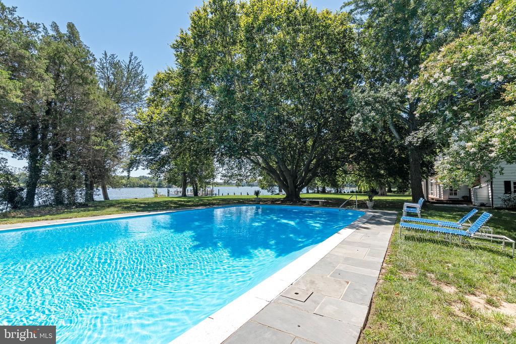 Swimming pool beside a stone patio, with lounge chairs, mature trees, and waterfront views.