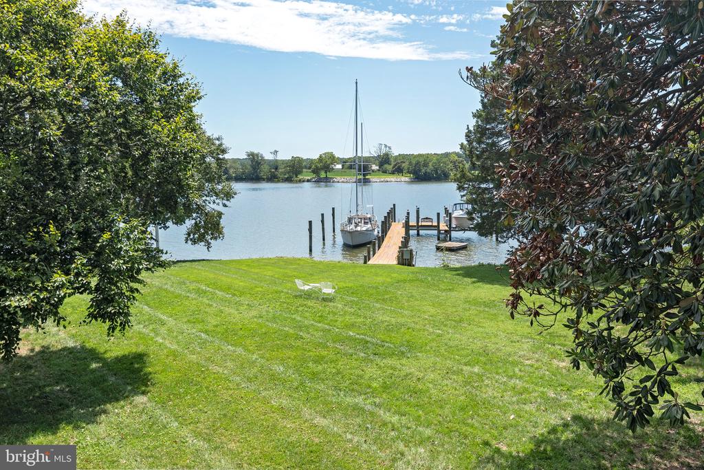 Waterfront lawn and dock with a moored sailboat, framed by mature trees.