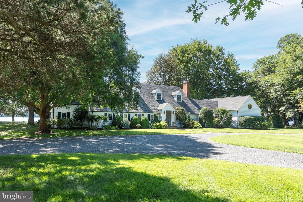 White house with dormer windows, a curving gravel driveway, and mature trees across the lawn.