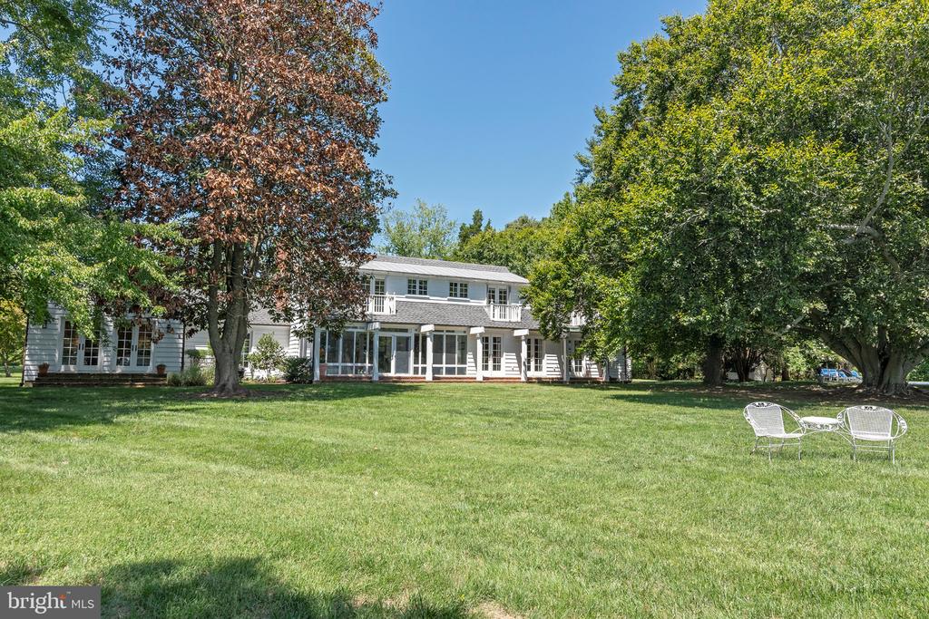 White two-story home beyond a broad lawn, framed by mature trees, with white patio chairs nearby.