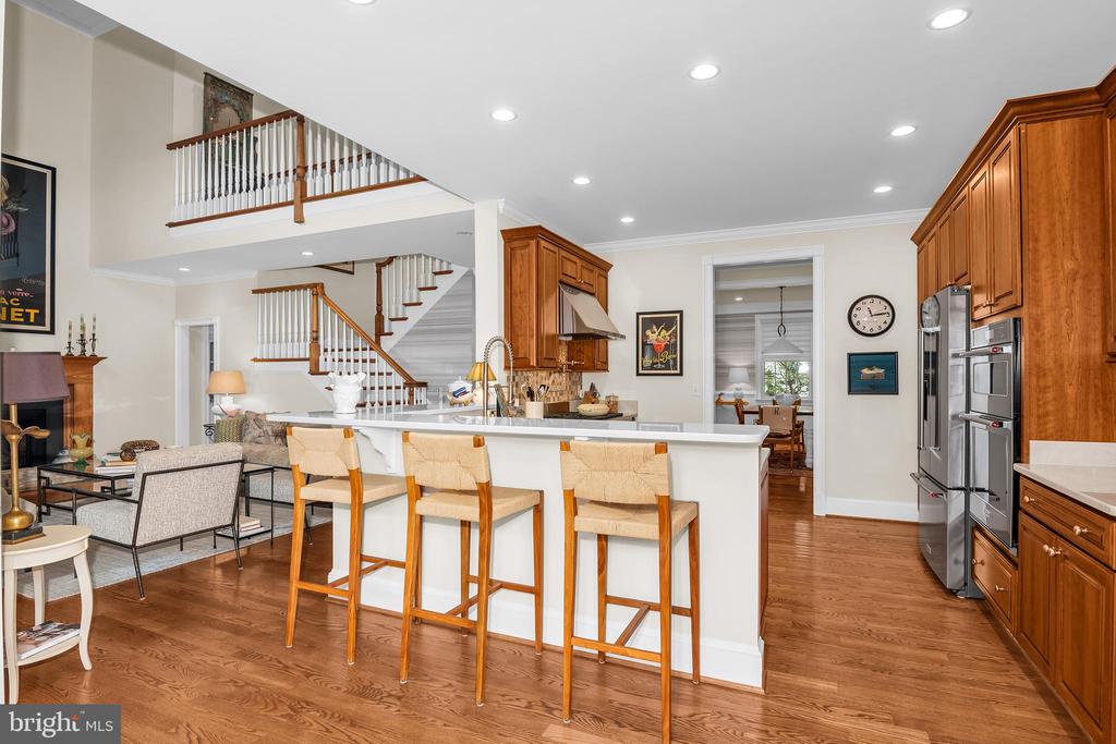 Open kitchen with a large white island, wood cabinetry, stainless appliances, and a staircase overlooking the living area.