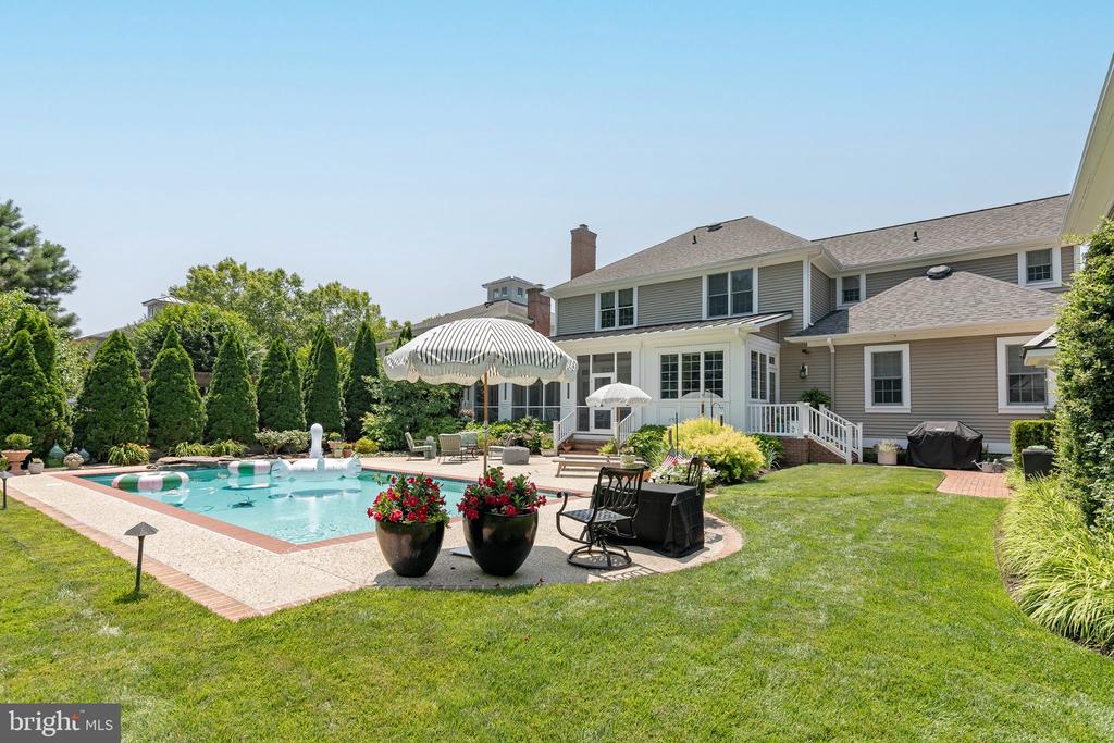 Rear of a two-story home with a swimming pool, striped umbrellas, patio furniture, and landscaped lawn.