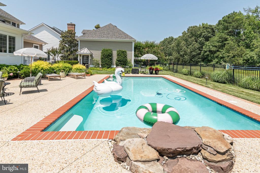 Swimming pool with a white swan float, stone patio, and fenced, tree-lined yard.