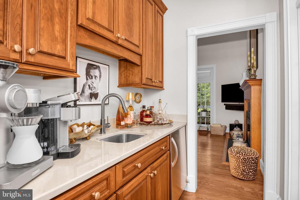 Kitchen beverage station with warm wood cabinets, a marble-look counter, and a small sink beside a living room doorway.