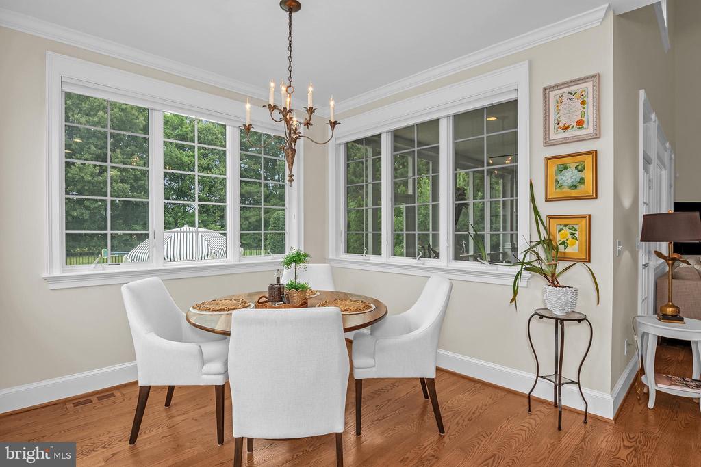 Dining room with a round table, white chairs, chandelier, and large windows overlooking leafy trees.