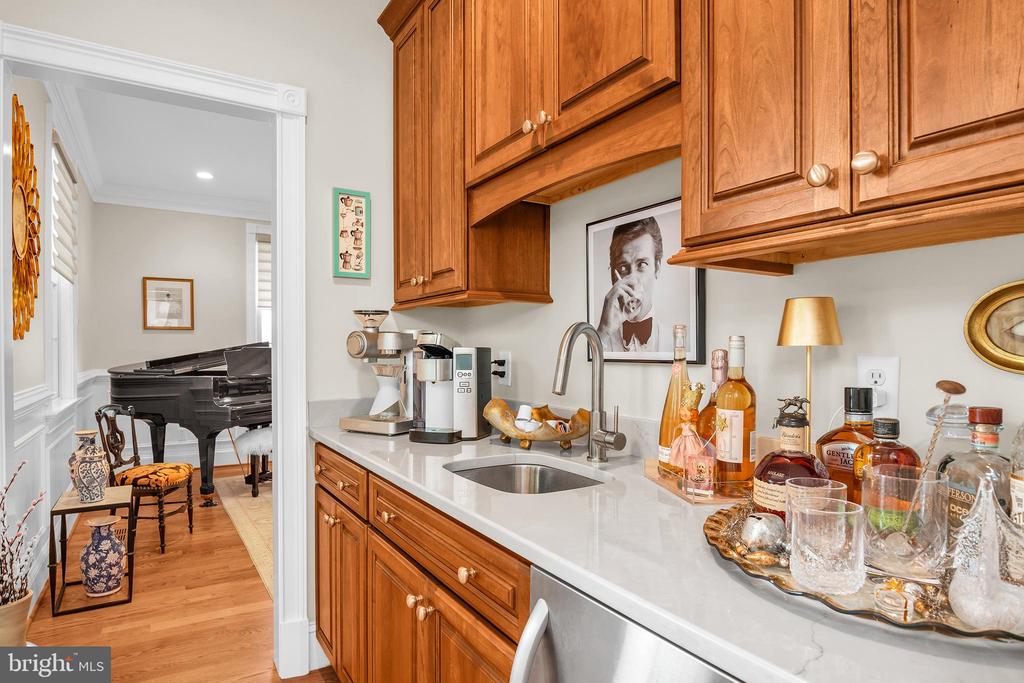 Kitchen with warm wood cabinetry and a white counter, opening onto a room with a grand piano.