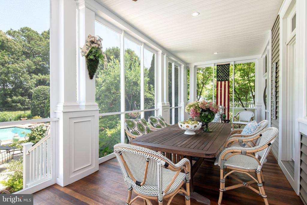 Screened-in porch with a wooden dining table, woven chairs, and views of trees and a swimming pool.