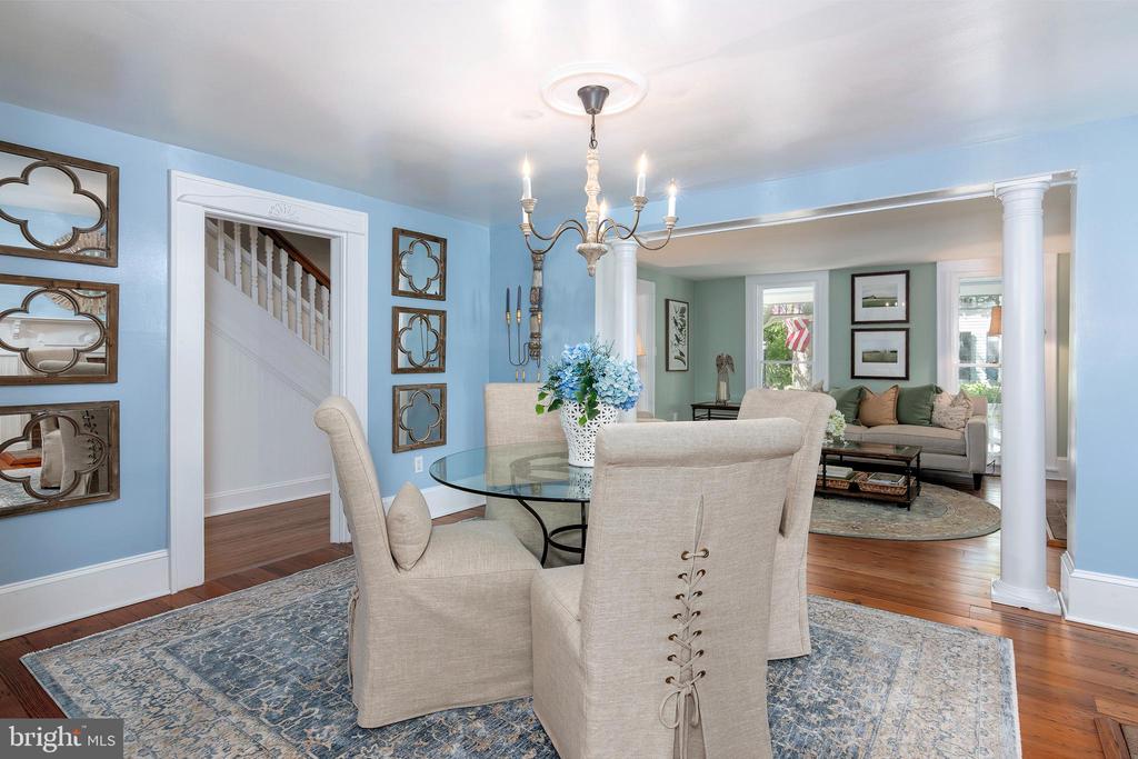 Dining room with a glass table and upholstered chairs opening into a living room with white columns.