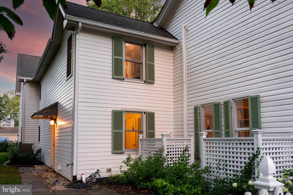 White-sided house with green shutters, glowing windows, and a white lattice fence at dusk.
