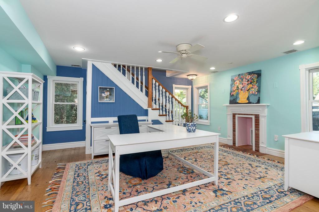 Home office with a white desk, blue chair, patterned rug, staircase, and small fireplace.