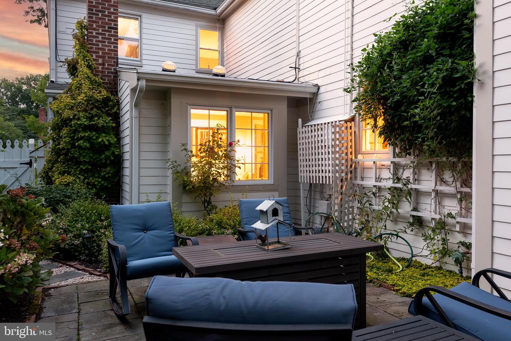Furnished stone patio with blue seating, garden beds, and warmly lit windows at dusk.