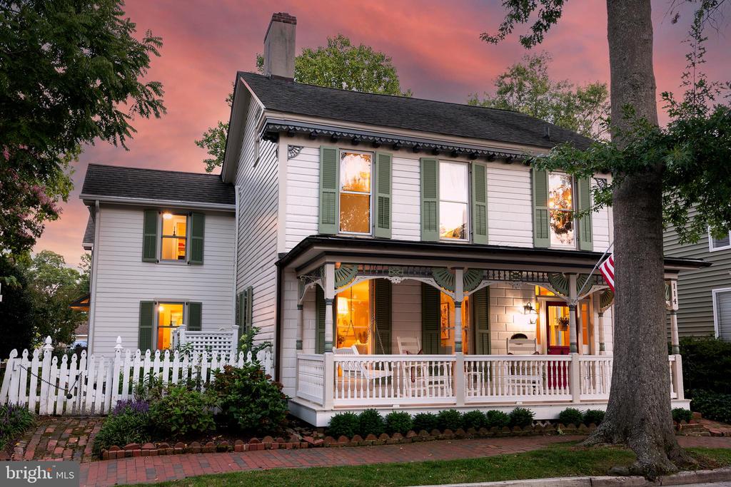 White two-story house with green shutters, a wraparound porch, and white picket fence at dusk.