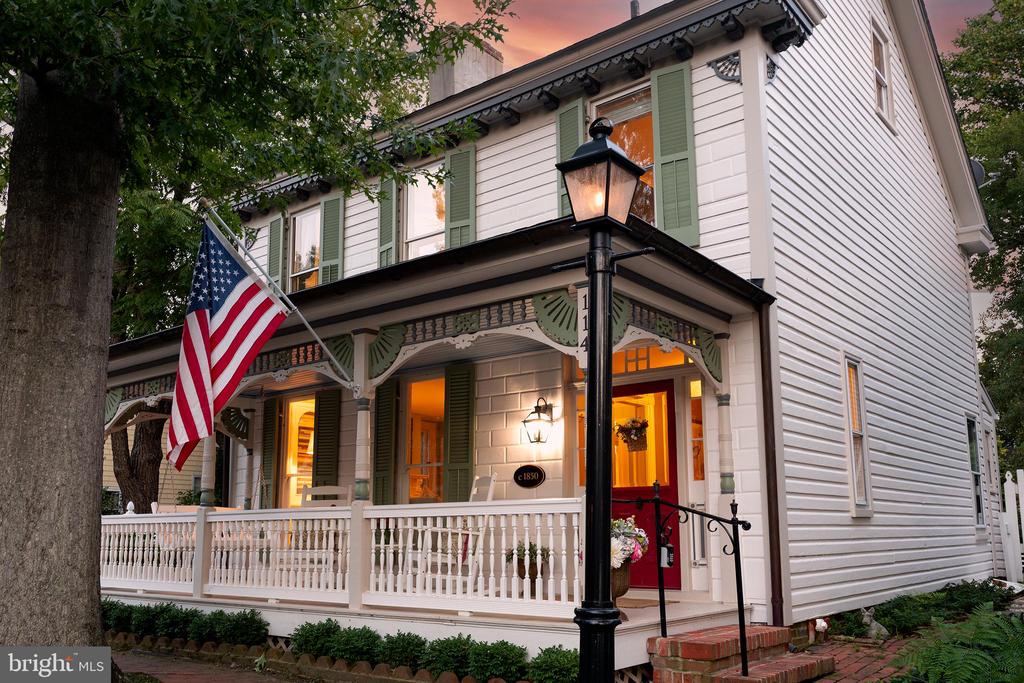Historic white clapboard house with green shutters, wraparound porch, American flag, and red front door.