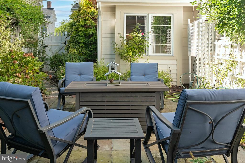 Patio with blue-cushioned chairs around a dark table, surrounded by leafy garden beds and white lattice fencing.