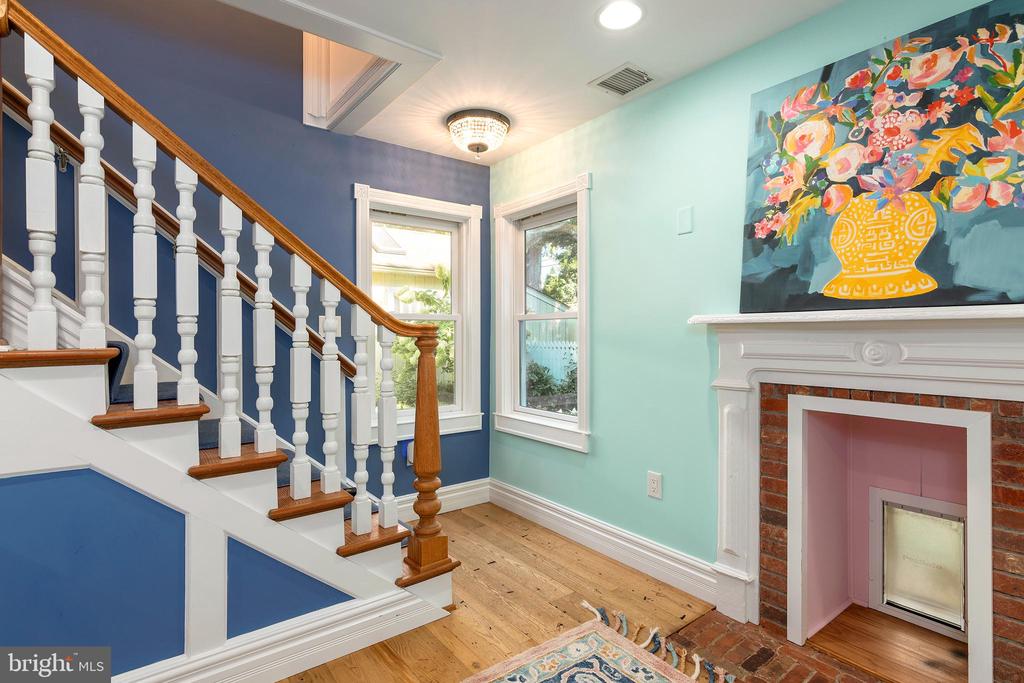 Bright entryway with a white staircase, polished wood railing, and brick fireplace beneath colorful floral artwork.