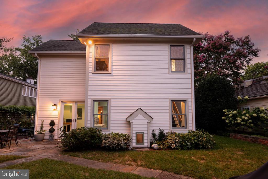 Rear of a white two-story house with a paved patio and garden beds at dusk.