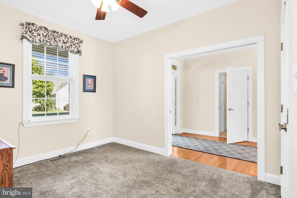 Empty carpeted room with a ceiling fan, floral window curtains, and a wide opening to the hardwood-floored hall.