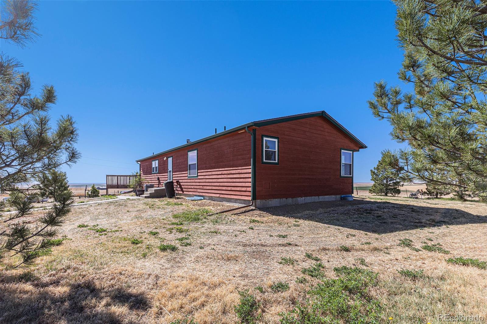 Red house with green trim in a dry yard, framed by pine trees and open farmland.