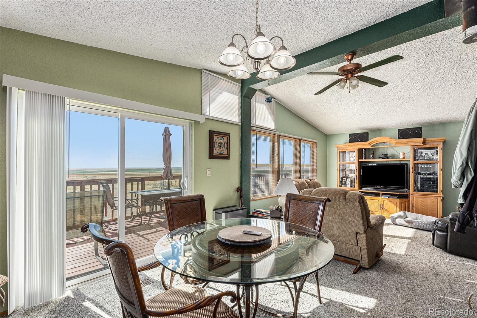 Living and dining room with vaulted ceiling, glass table, and sliding doors to a deck overlooking open fields.