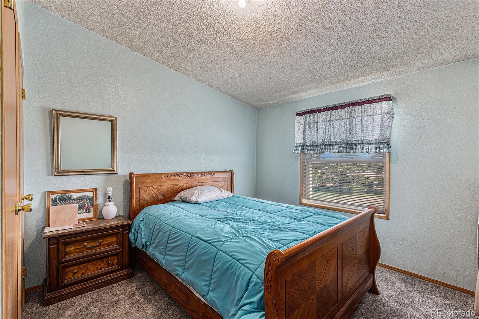 Bedroom with a carved wooden bed, turquoise quilt, matching nightstand, and curtained window.