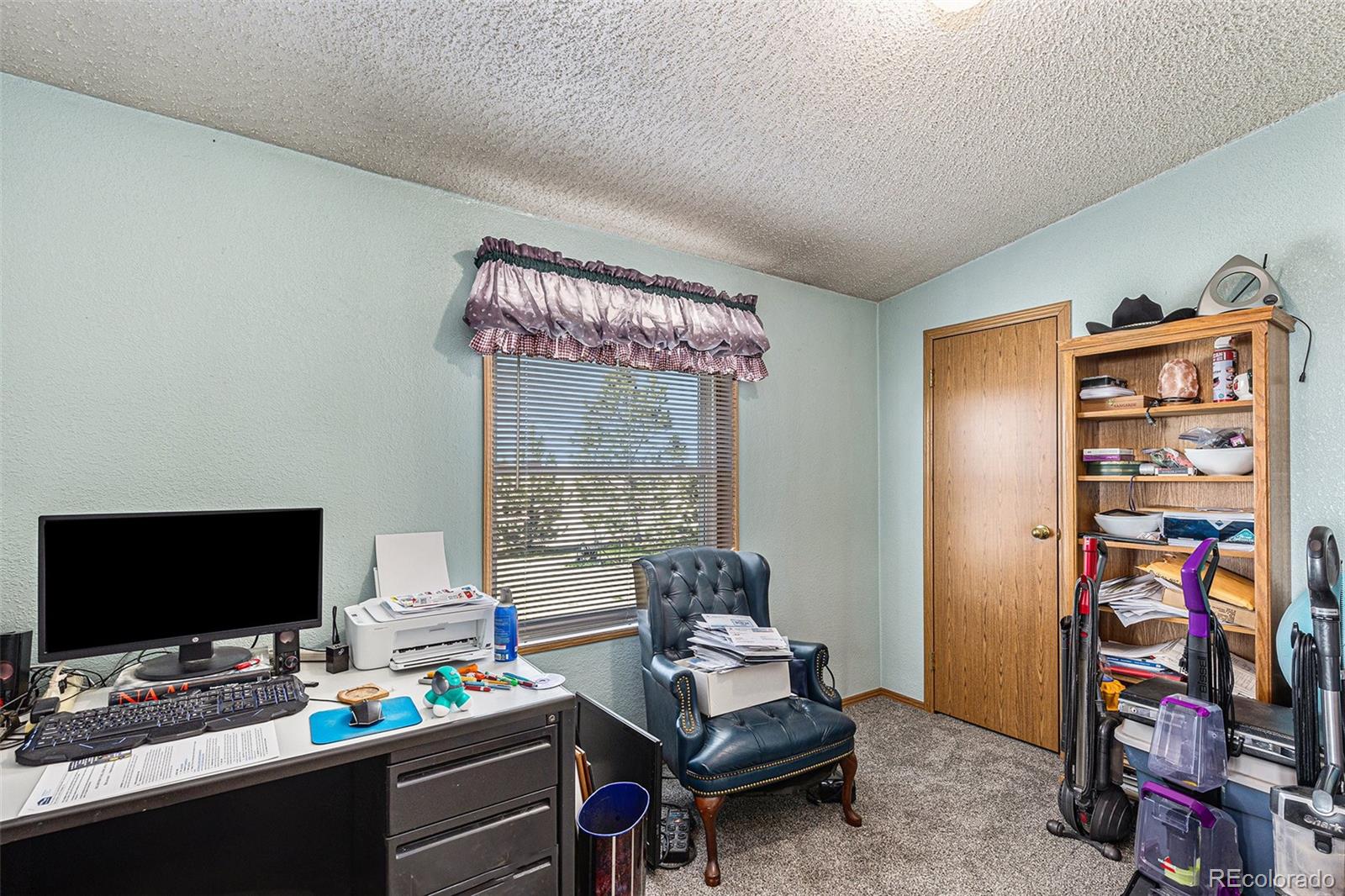Home office with a computer desk, leather chair, window blinds, and shelves beside several vacuum cleaners.