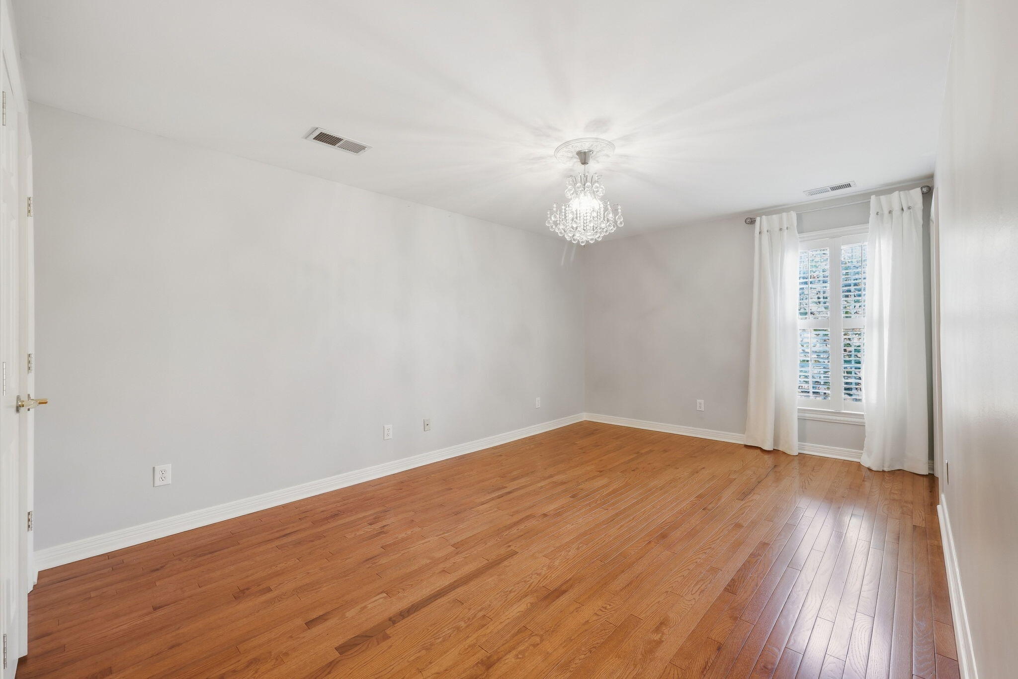 Empty room with hardwood floors, a crystal chandelier, and a curtained shuttered window.