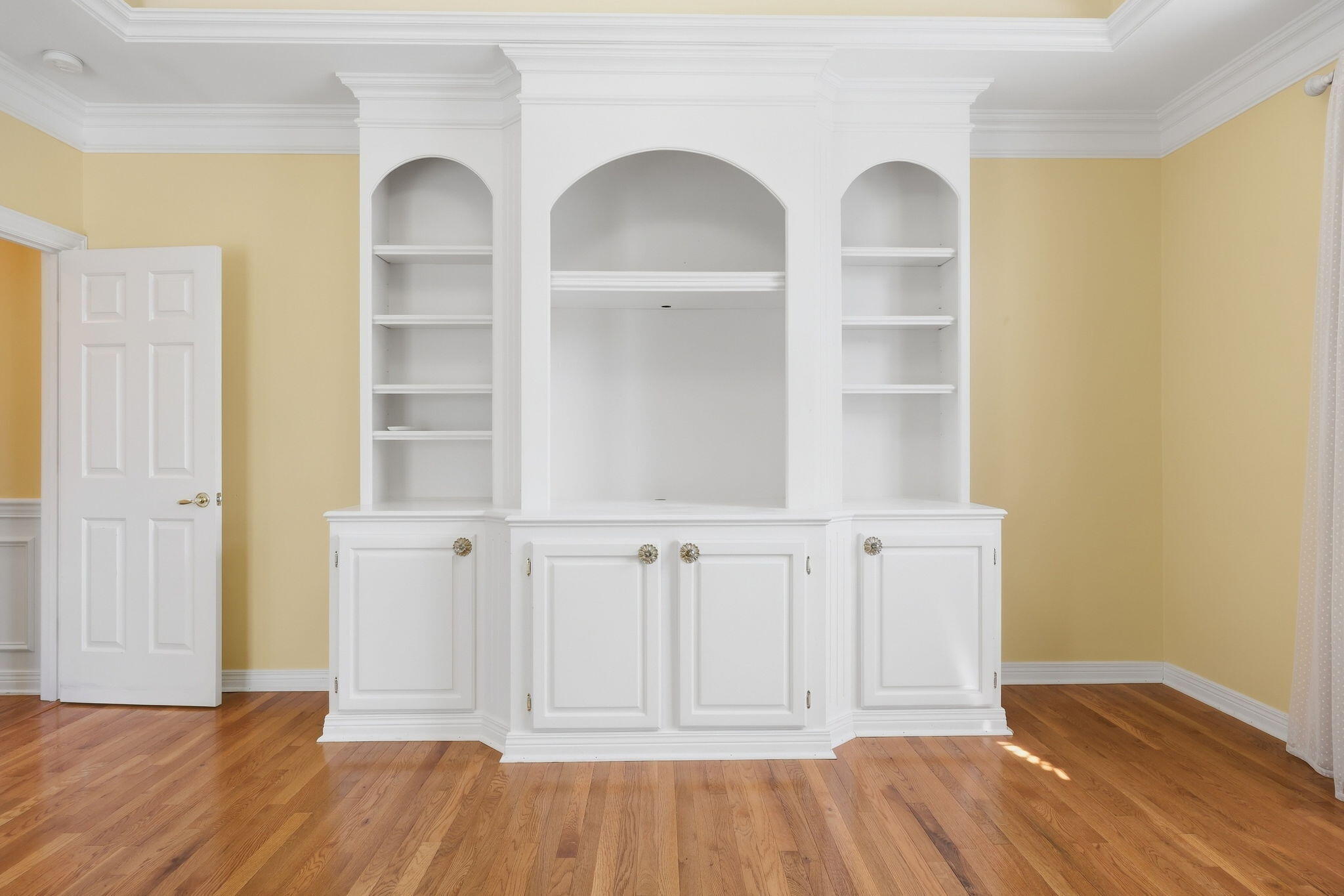 Empty room with a white built-in cabinet, arched shelves, and hardwood flooring.