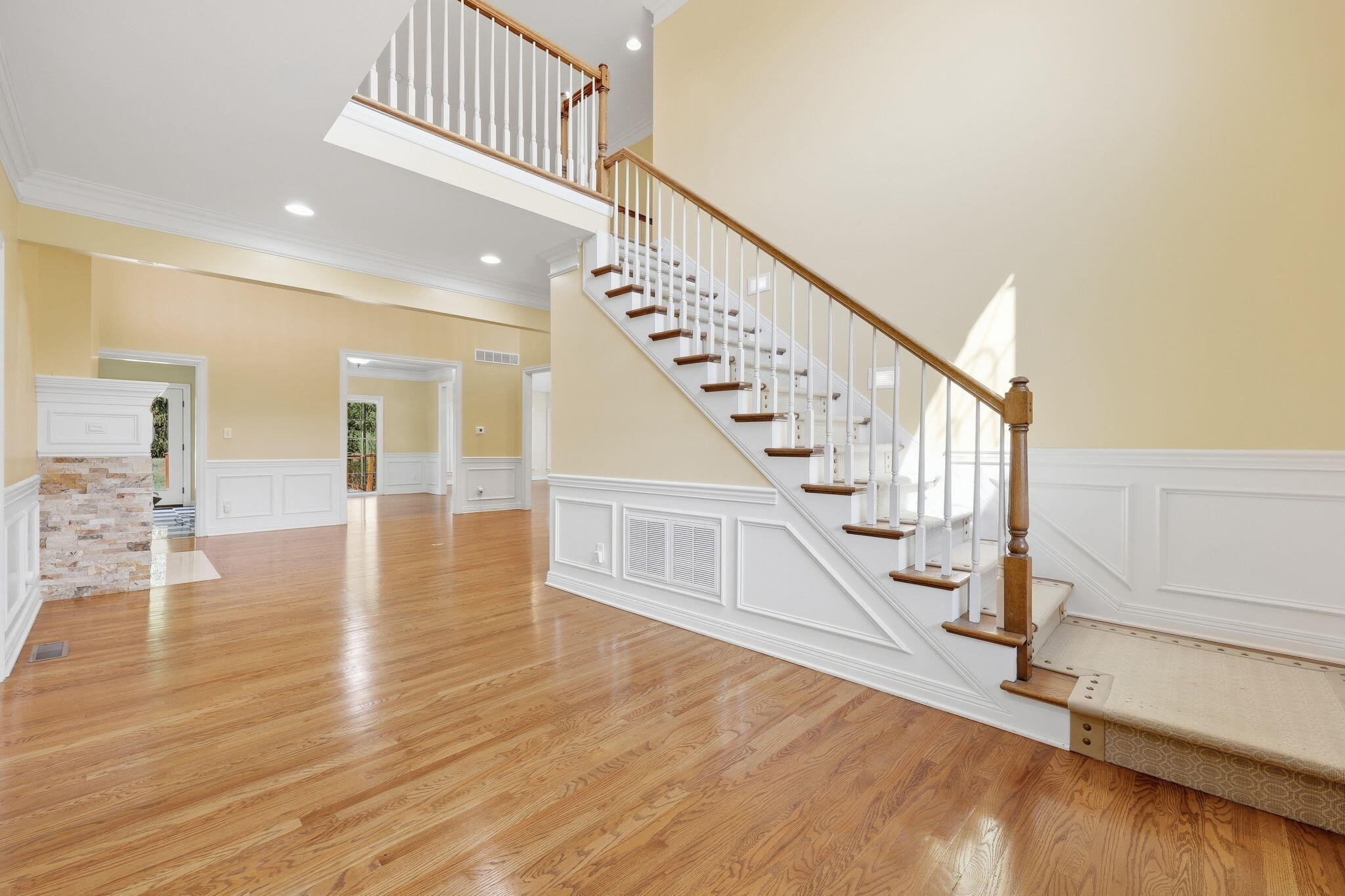 Two-story foyer with polished hardwood floors, white-trimmed staircase, and a stone fireplace visible through a doorway.