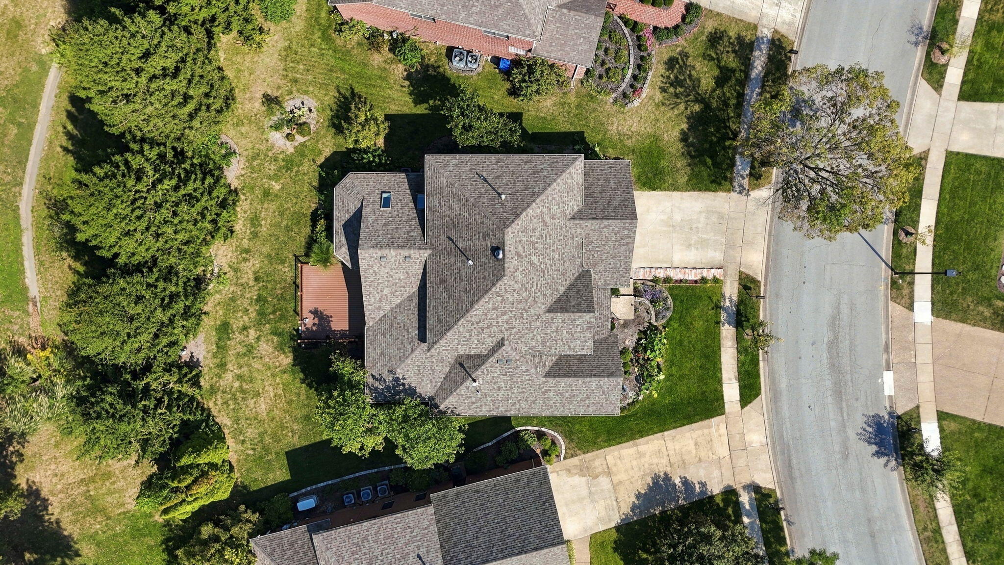 Aerial view of a gray-roofed home with a wooden deck, landscaped lawn, and mature trees.