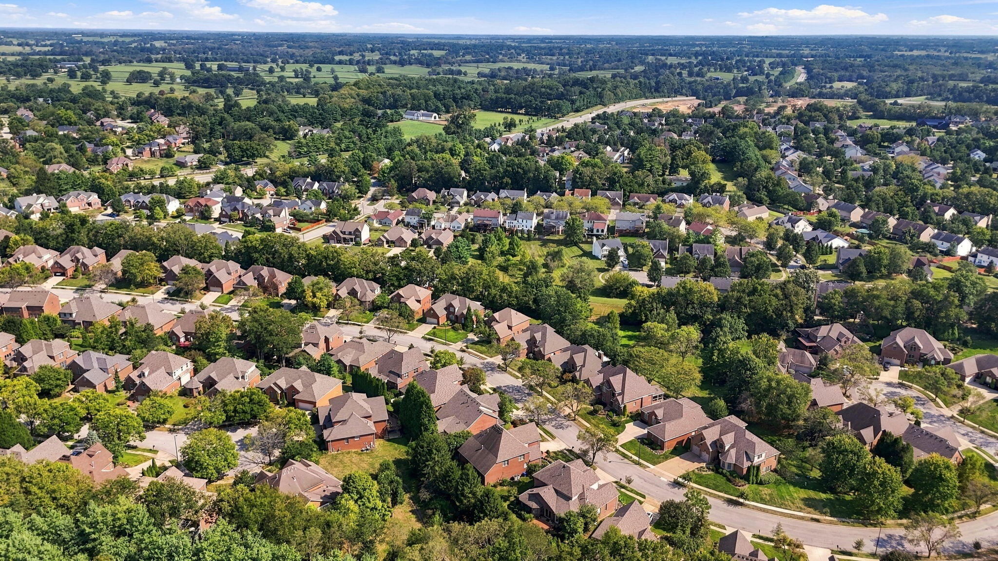 Aerial view of a leafy neighborhood with detached homes, tree-lined streets, and open fields beyond.