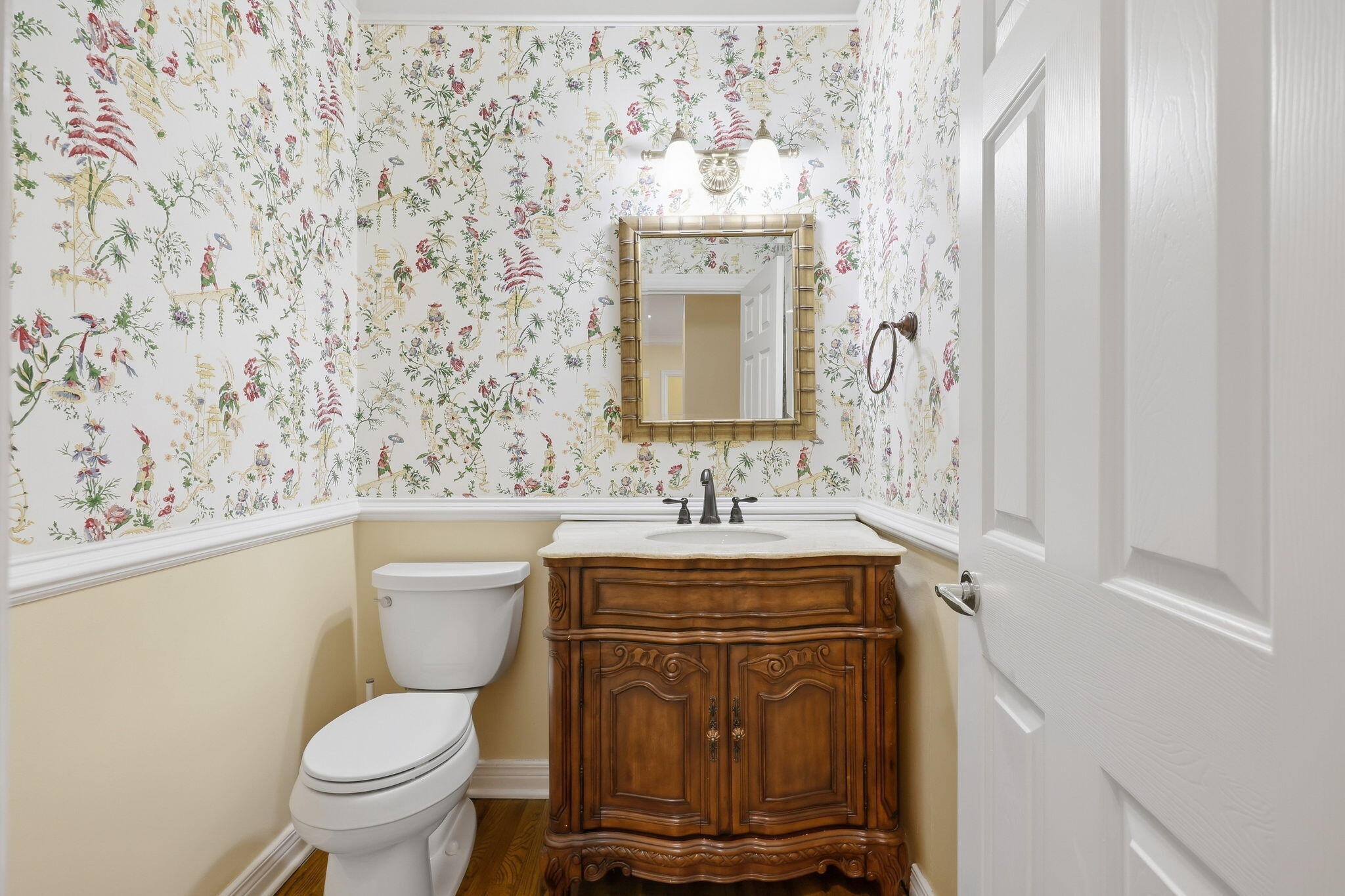 Powder room with an ornate dark wood vanity, white toilet, and colorful floral wallpaper.