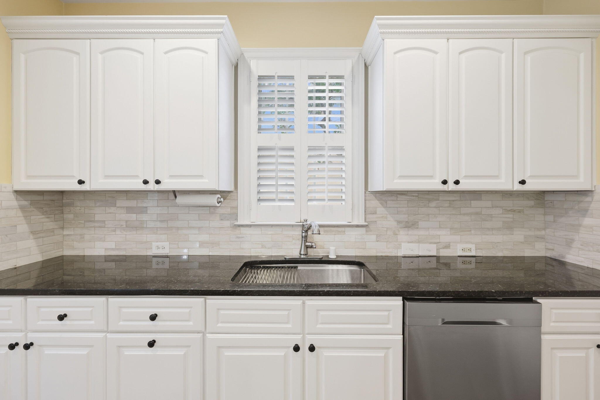 Kitchen with white cabinets, black countertops, a stainless steel sink, and plantation shutters.