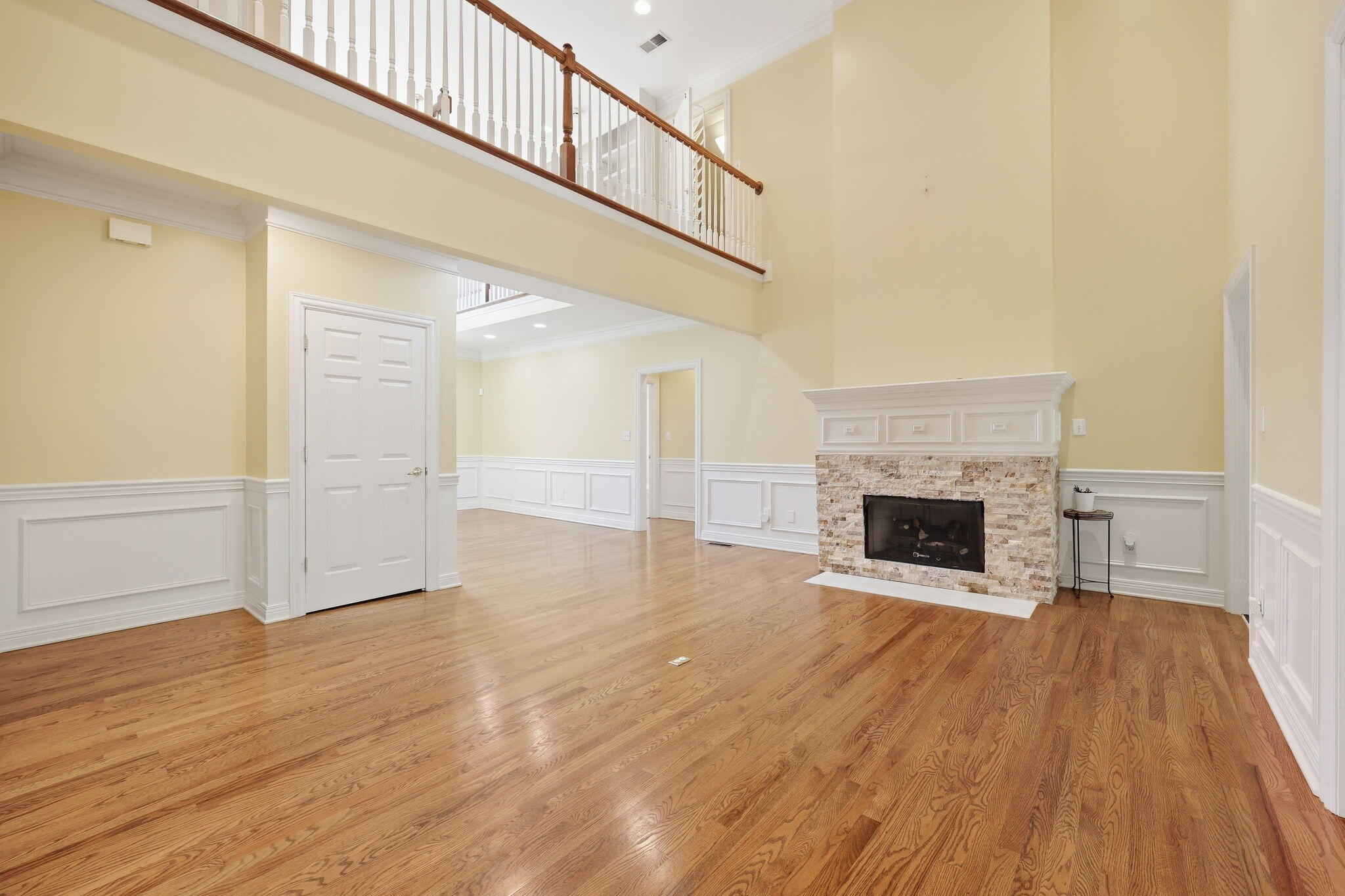 Empty two-story living room with hardwood floors, a stone fireplace, and an upstairs balcony.