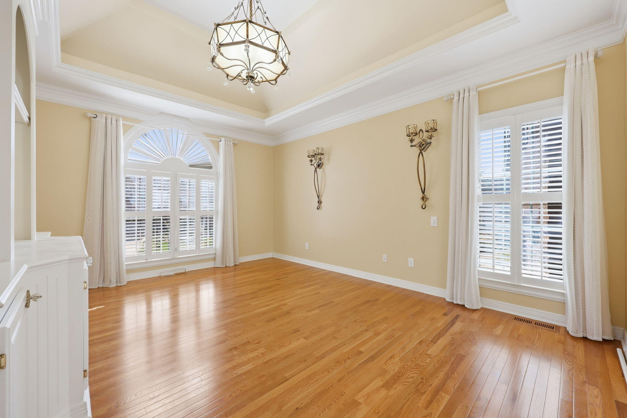 Empty dining room with hardwood floors, an ornate chandelier, and tall shuttered windows, one with an arched top.