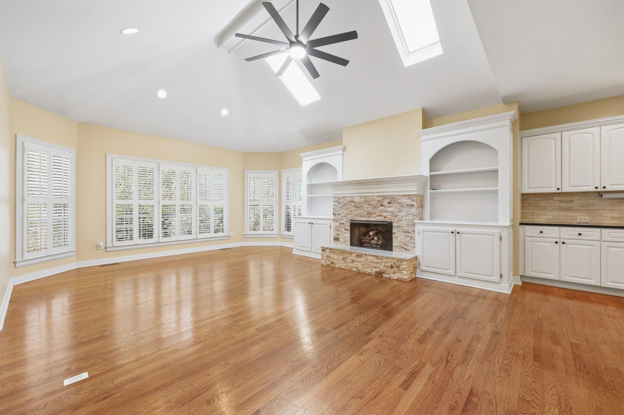 Empty living room with hardwood floors, stone fireplace, white built-ins, shuttered windows, and vaulted skylit ceiling.