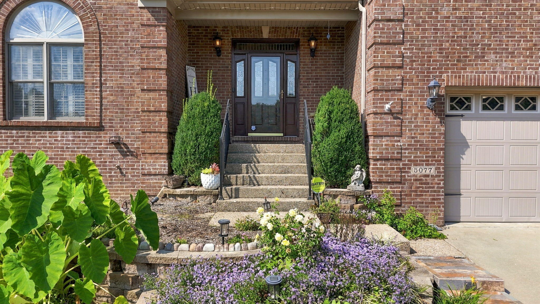 Red-brick home entrance with concrete steps, flowering garden beds, and an attached garage.