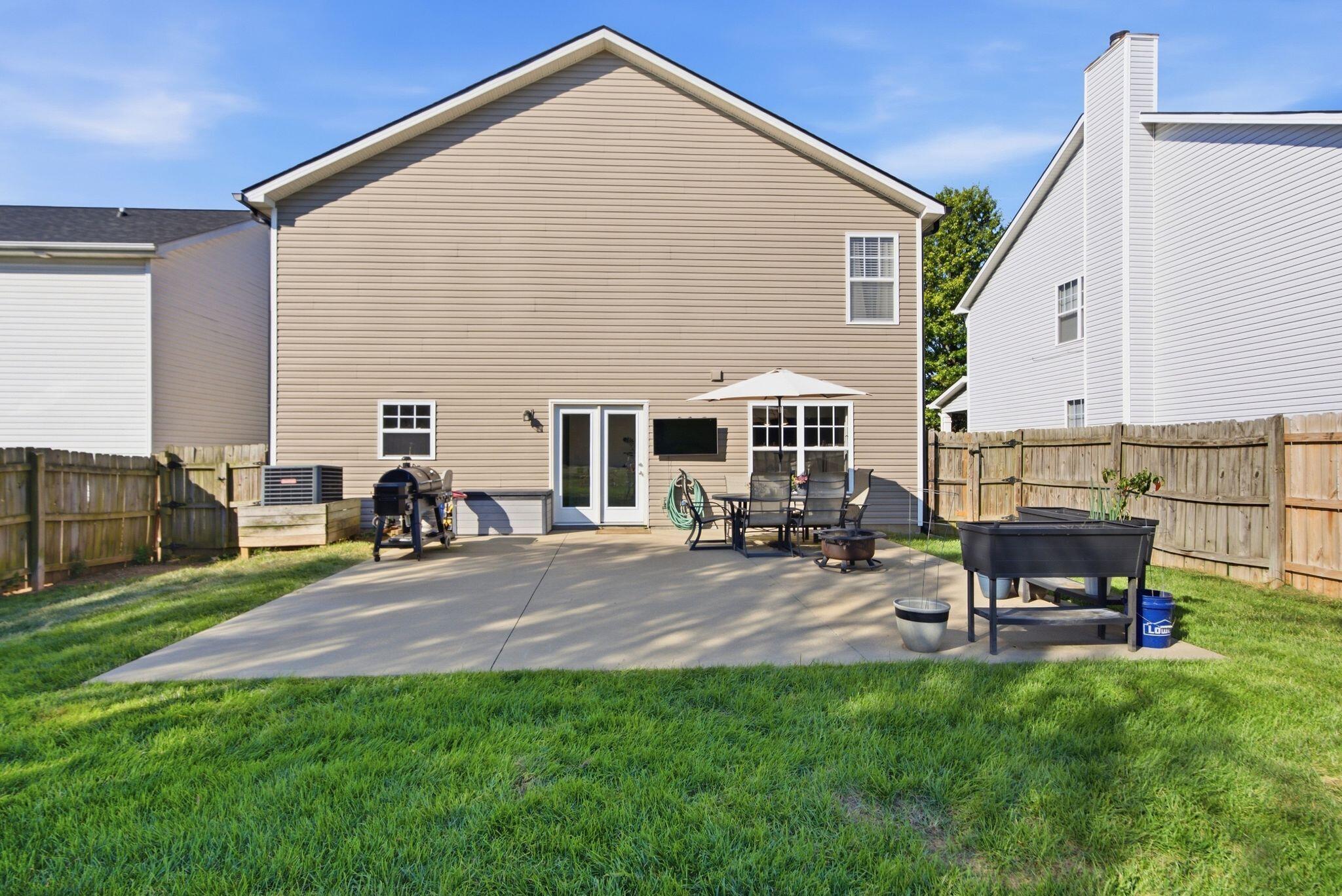 Back of a tan-sided home with a fenced backyard patio, outdoor dining set, umbrella, and grill.