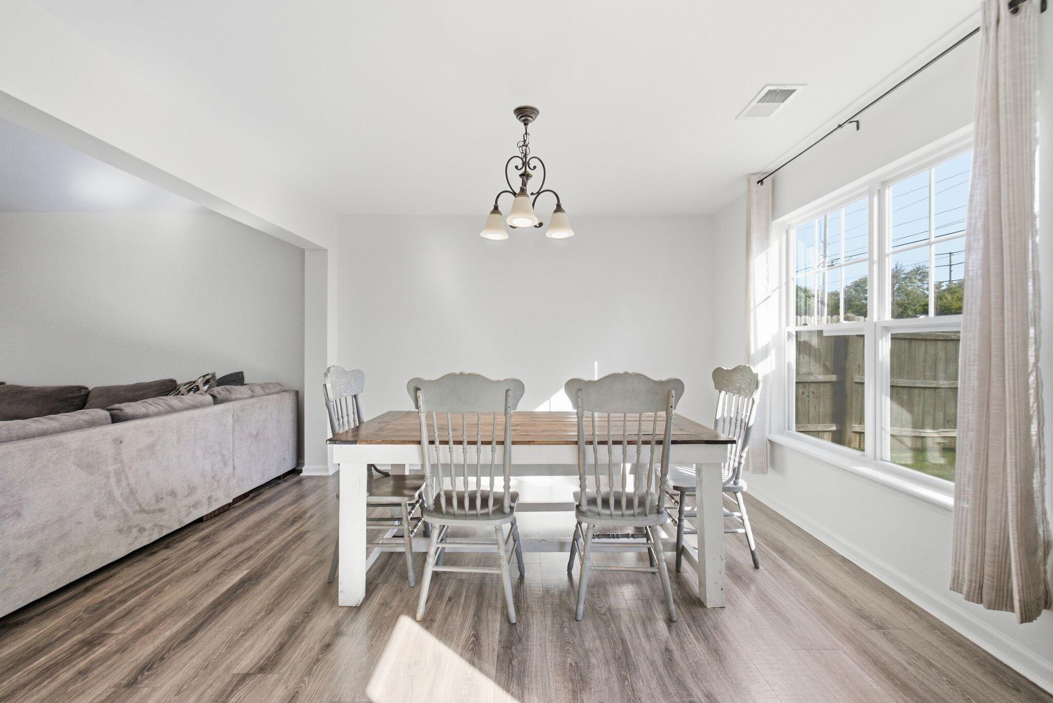 Dining area with a wood-top table and white chairs beside large sunlit windows, opening to a living area.