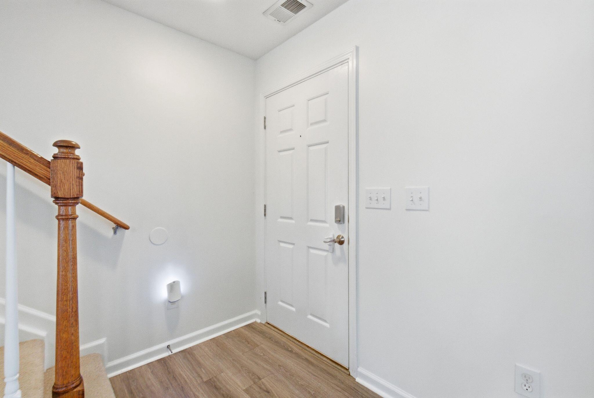 Entry foyer with a white paneled front door, wood-look flooring, and a wooden staircase banister.