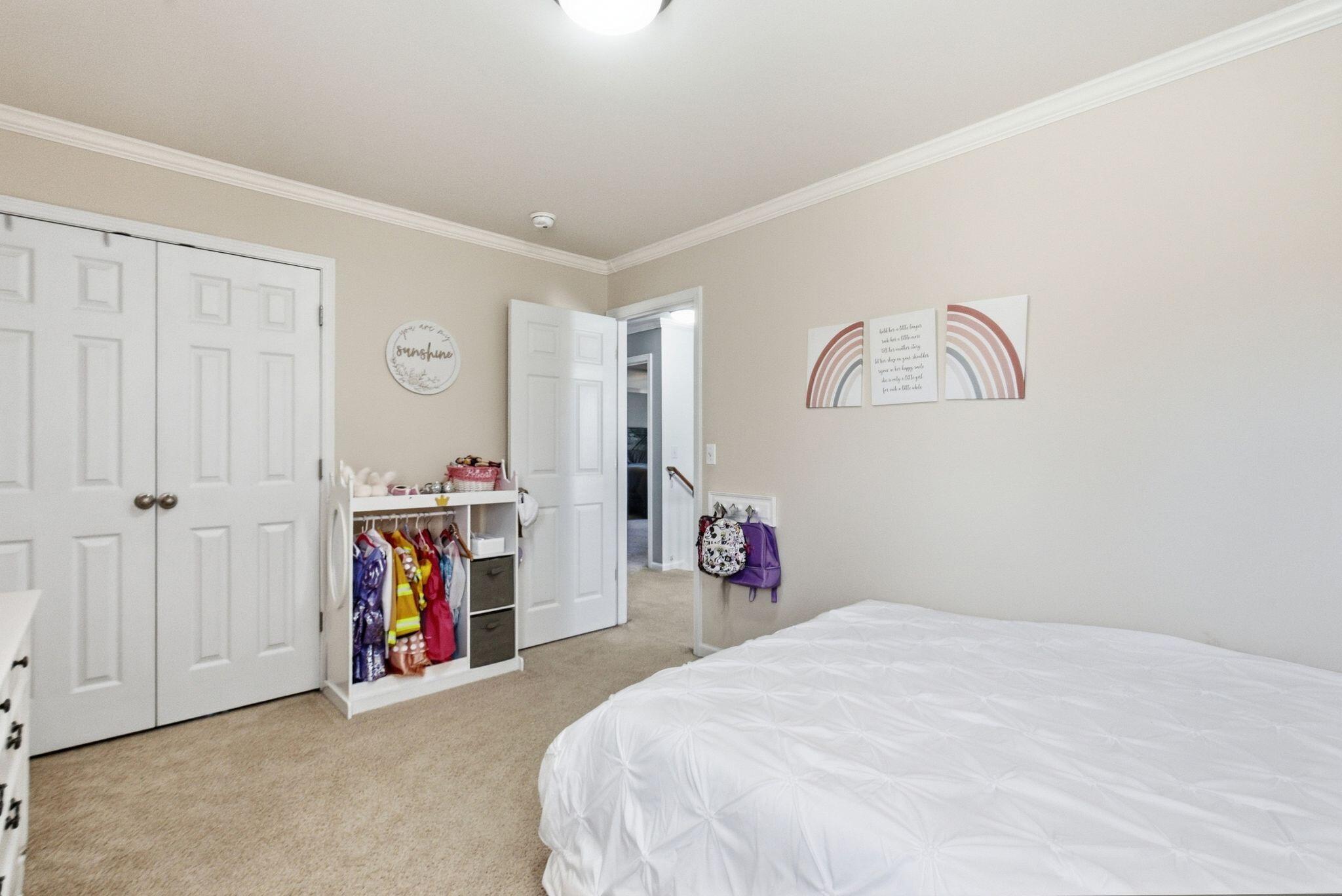 Bedroom with a white bed, beige carpet, closet doors, and a clothing rack with colorful garments.