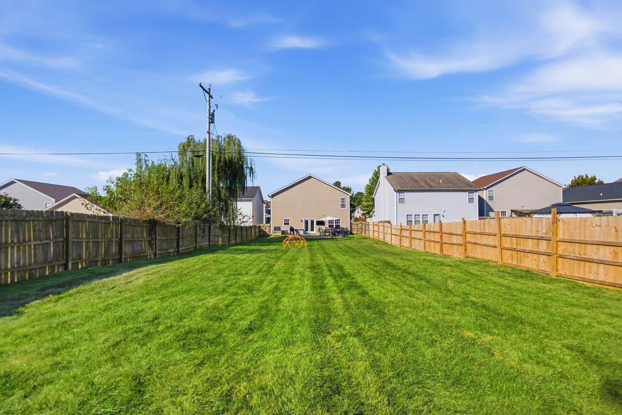 Fenced backyard with a broad green lawn, small play structure, and patio furniture near the house.