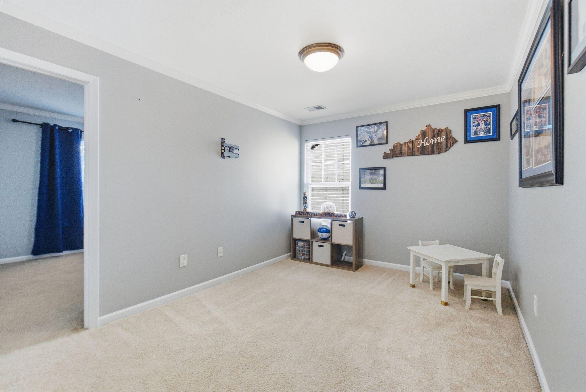 Carpeted bonus room with a small table, sports memorabilia, and a doorway to a room with blue curtains.