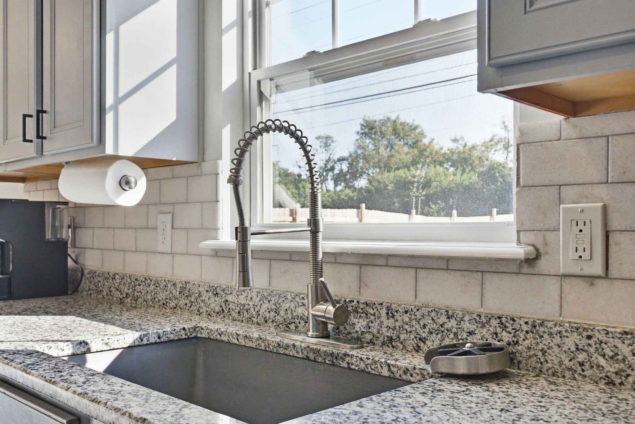 Kitchen sink with a spring-neck faucet, granite counters, white tile backsplash, and sunlit window.