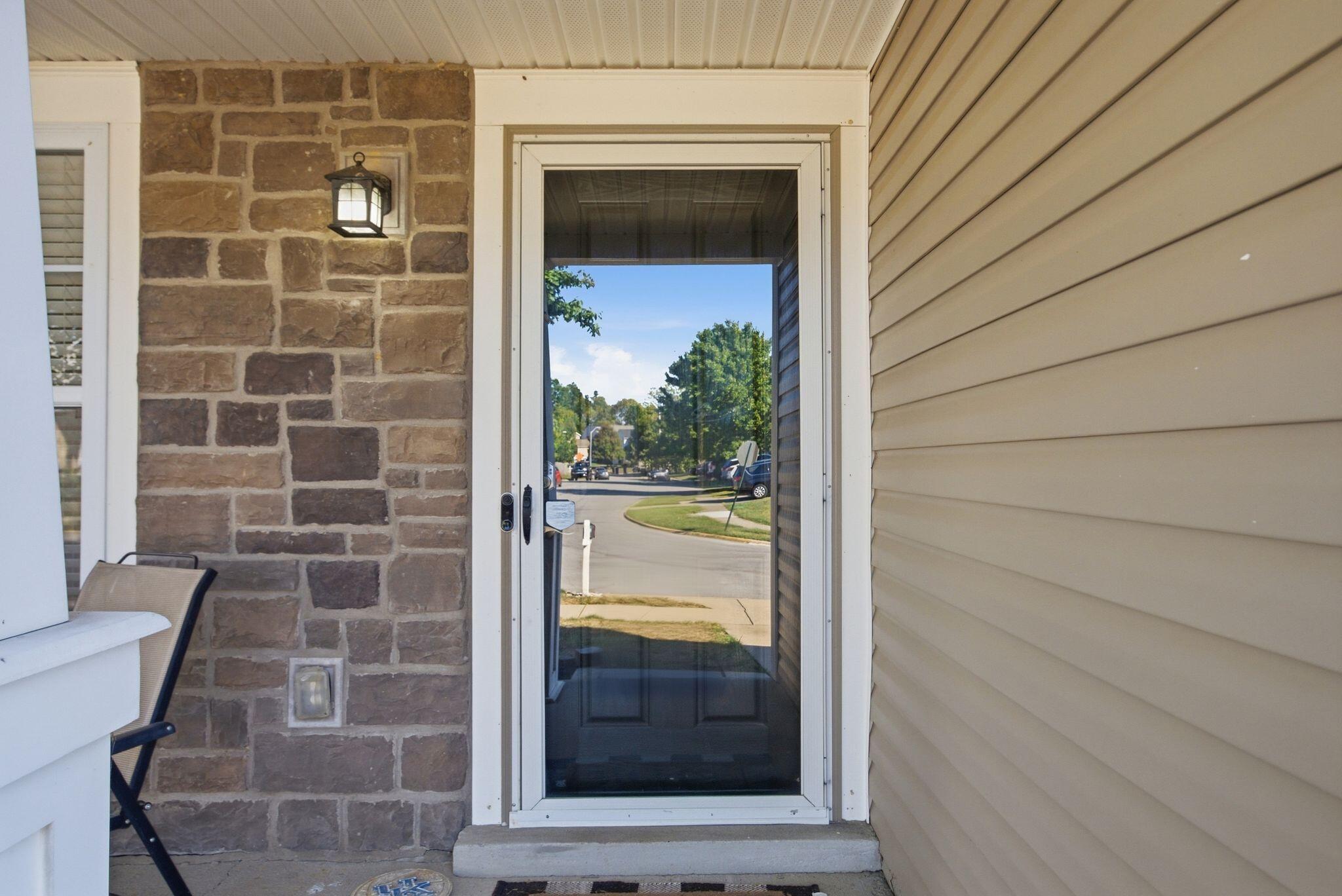Covered front entry with a glass storm door, stone accent wall, and tan siding.